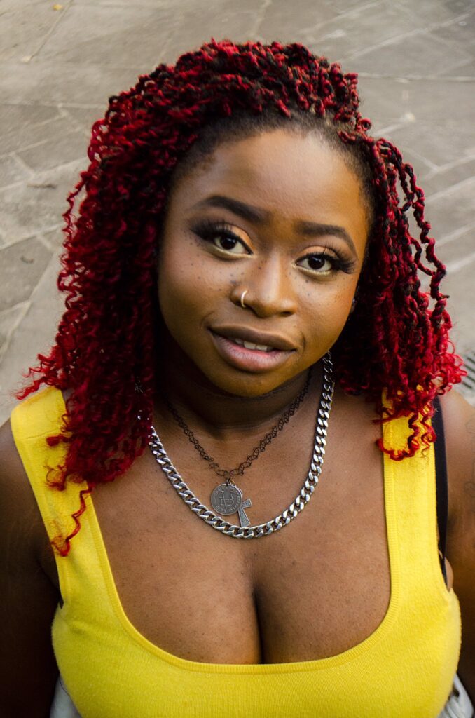 A woman of colour with vibrant red hair looks directly at the camera, which appears elevated above her. She wears a yellow tank top, a nose ring on her right nostril, and two necklaces—one with an ankh and medallion pendant, the other a bold silver Cuban link chain.