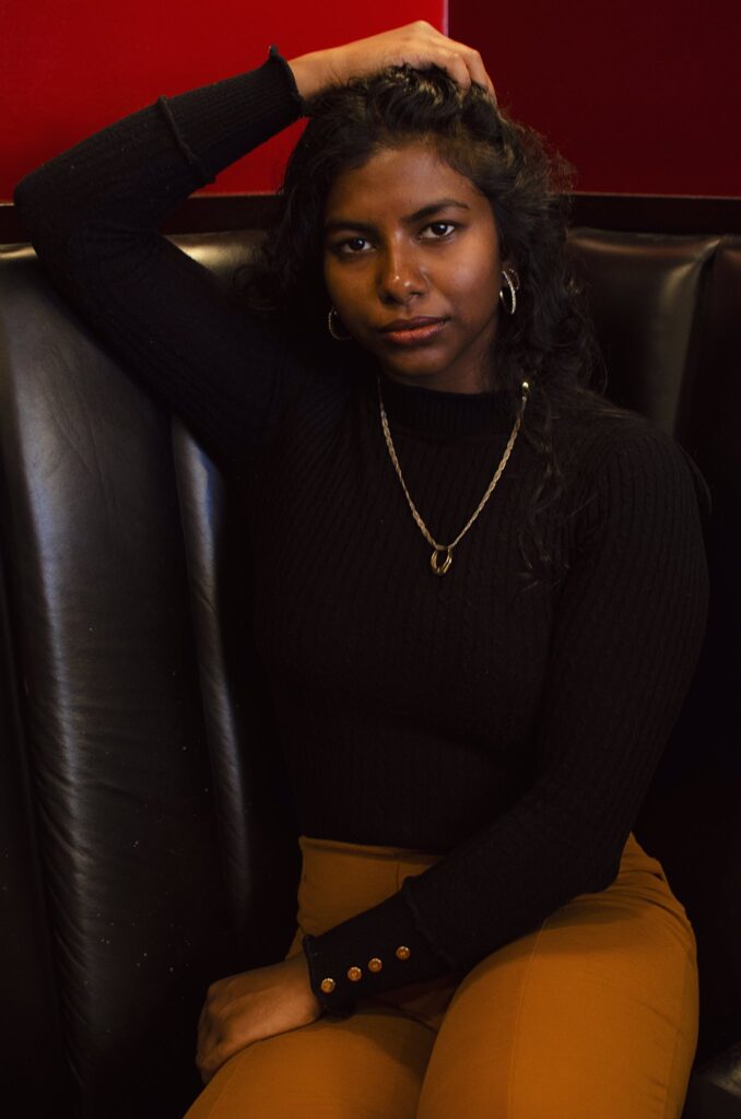 A coloured woman sitting at a booth, one hand running through her hair while the other rests on her lap, gazing directly at the camera. She wears a gold necklace, a black turtleneck, and brown pants, exuding a relaxed yet confident vibe.