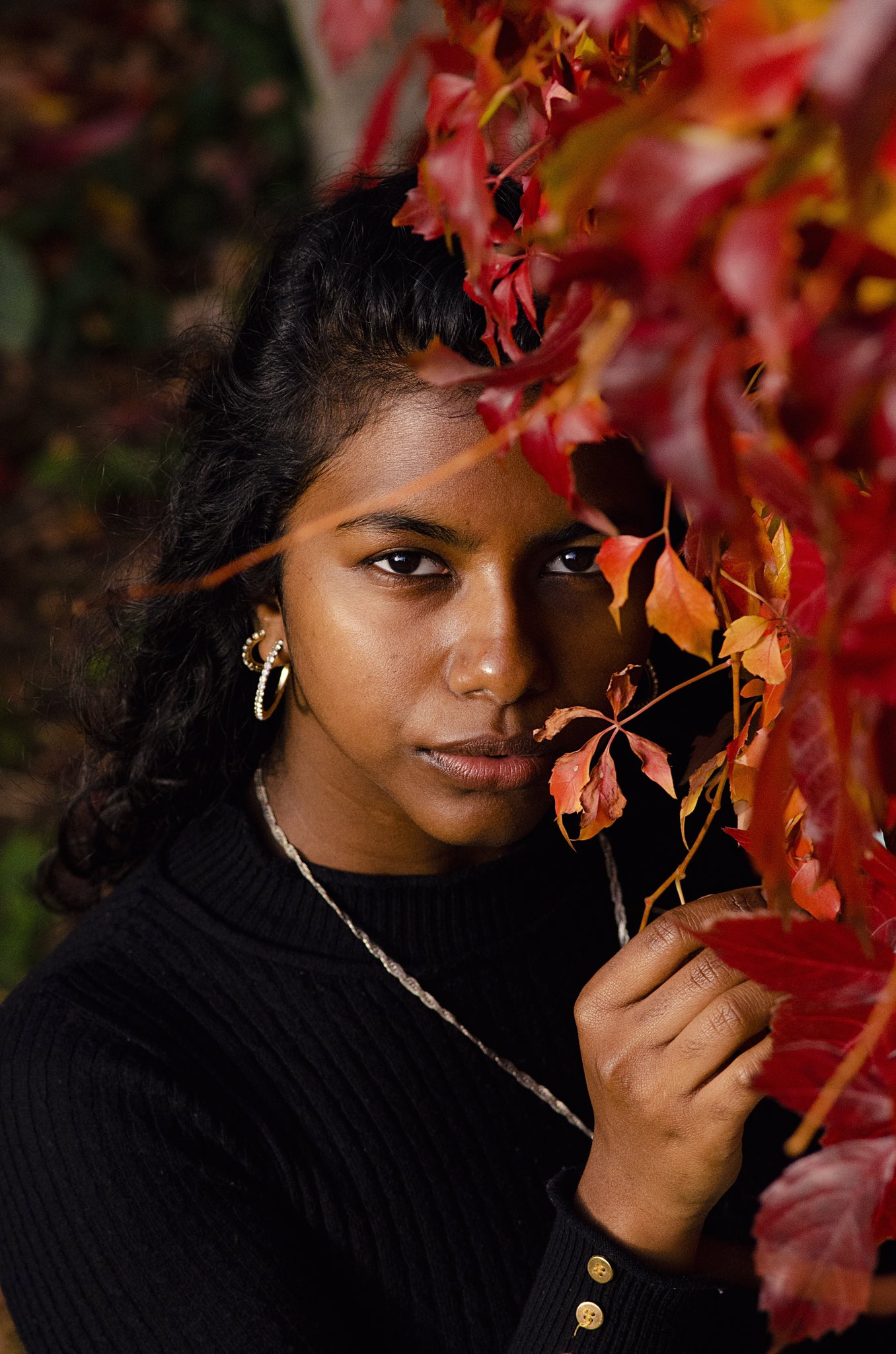 A close-up of a woman of colour peeking through vibrant red leaves, one hand gently resting on them as she looks directly at the camera, her expression both curious and engaging.