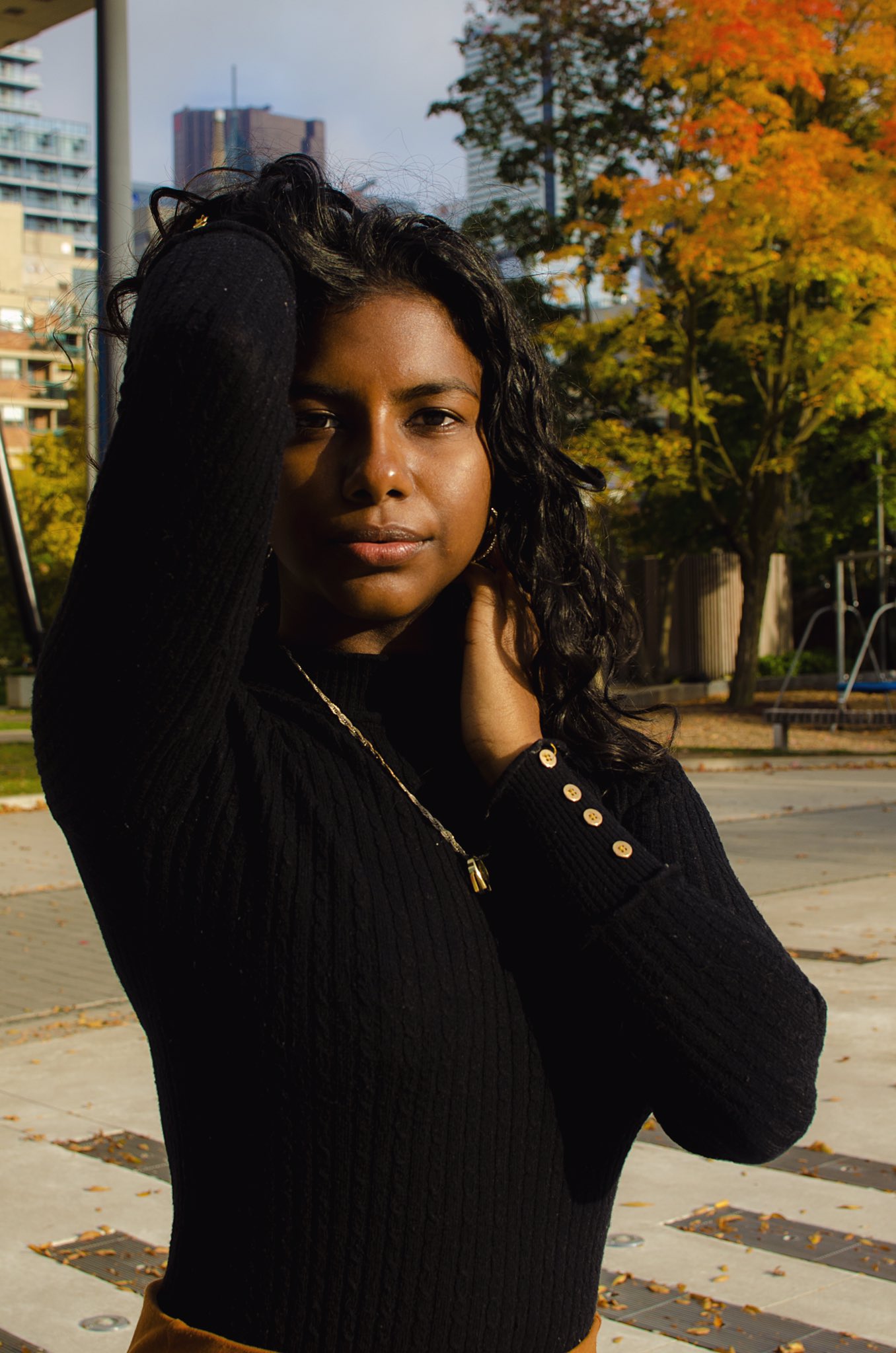 A woman of colour poses outdoors, one arm lifted and extended backward, while the other gently rests on her cheek as she gazes directly at the camera.
