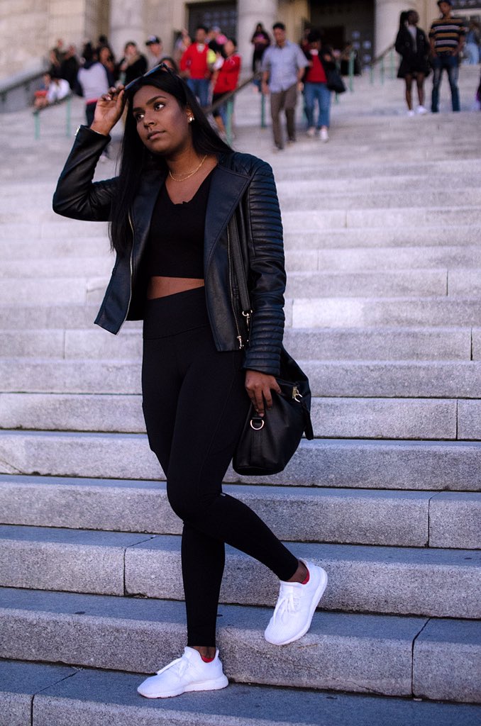 A candid image of a woman of colour walking down some steps. She has her hands on her glasses, which are positioned on top of her head. She is wearing a full black outfit with part of her belly showing in a stylish way and white contrasting shoes.