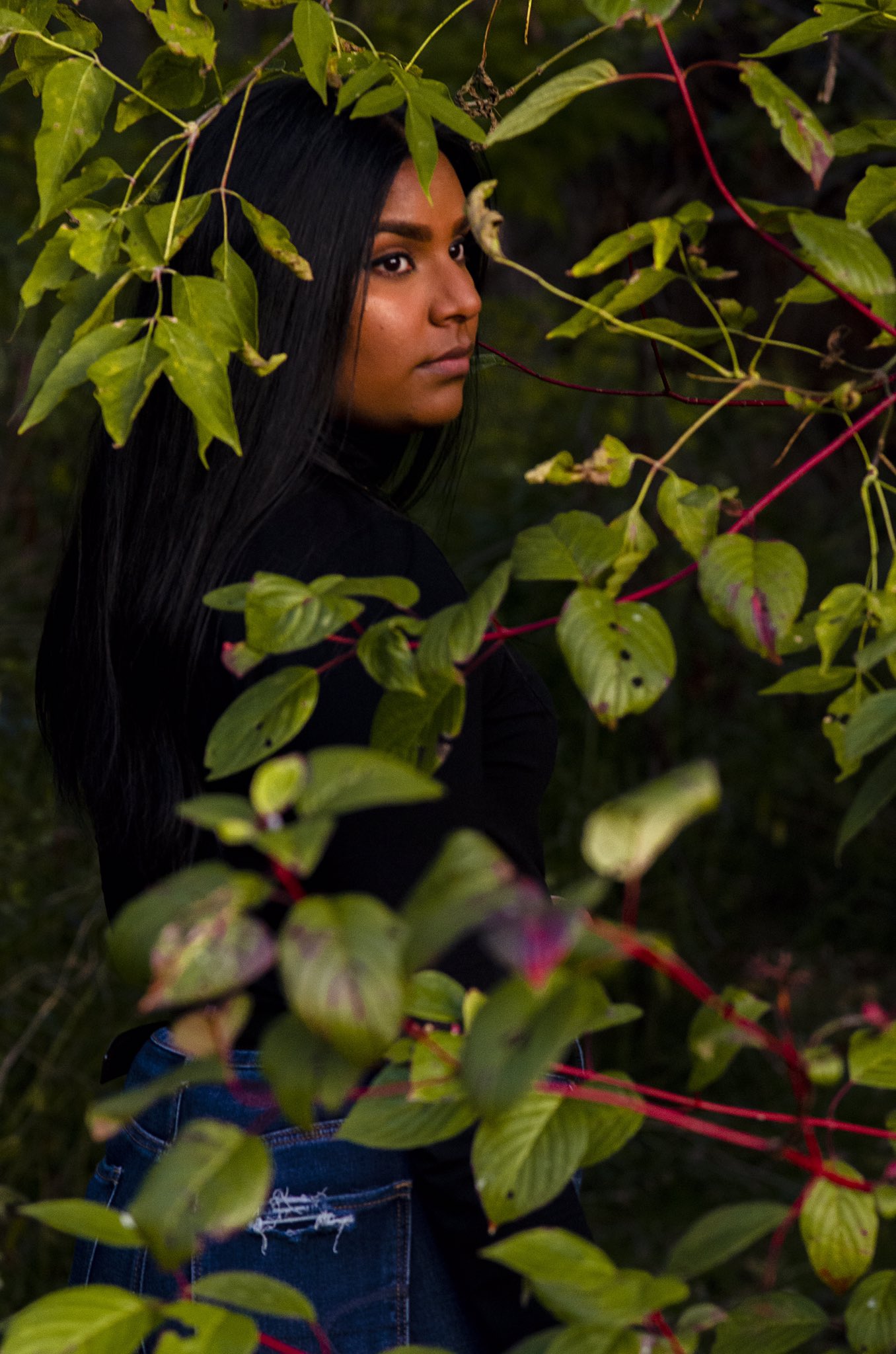 A woman of colour stands with her back to the camera in a forested area, looking back over her shoulder at about a 45-degree angle.