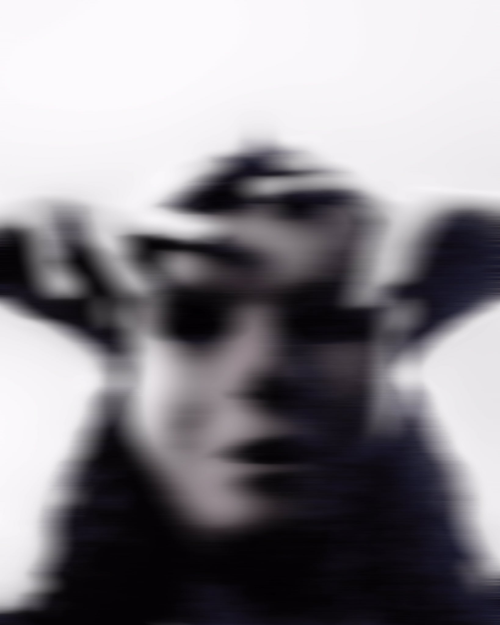 A blurred black and white headshot of a masked individual, with shadowed eye sockets that evoke mystery and anonymity. Their hands rest on the crown of their head, enhancing the sense of tension and intrigue.