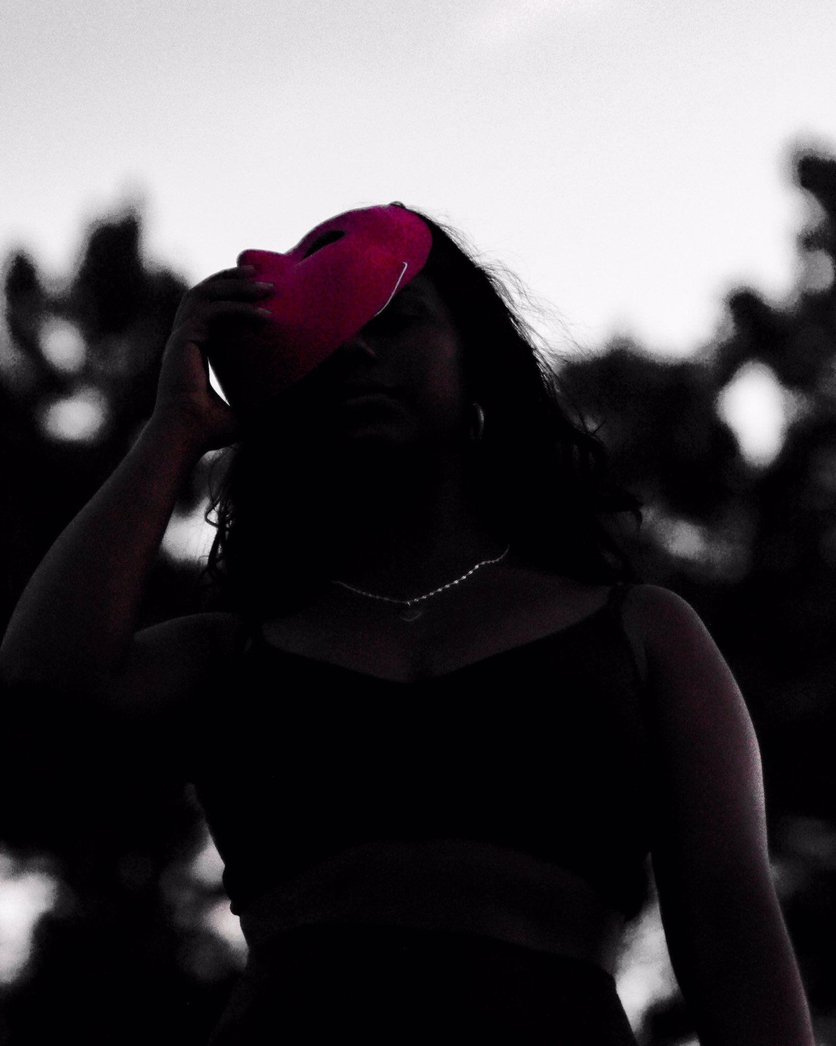 A woman in a black dress and pearl necklace stands against a pale, overexposed sky, captured mid-motion as she removes a vibrant red mask—creating a striking contrast of color and emotion.