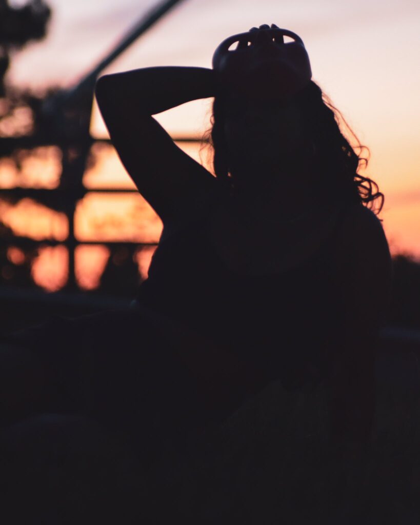 A warm silhouette of a woman gently removing a mask, its outline faint against the glowing hues of a sunset backdrop—evoking themes of transformation and quiet release.